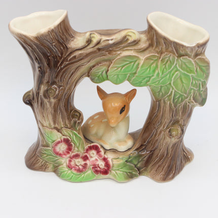 A ceramic double candle holder designed to look like a tree stump, featuring a small fawn figurine in the center, green leaves, and pink flowers on a white background.