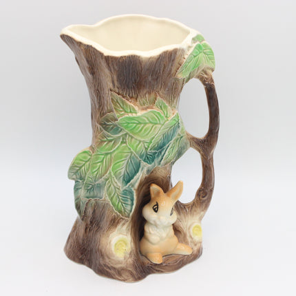 A ceramic pitcher shaped like a tree trunk with green leaves, a small rabbit figurine in a hollow at the base, and a cream-colored interior, shown on a light gray background.