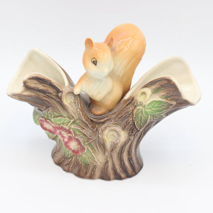 A ceramic figurine of a brown and cream squirrel perched on a tree trunk base with painted flowers and leaves, photographed on a plain white background.
