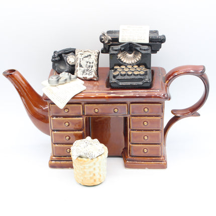 A brown ceramic teapot shaped like a vintage desk, decorated with miniature black typewriter, rotary phone, and silver paperclips, with a small woven basket in front, set against a plain white background.