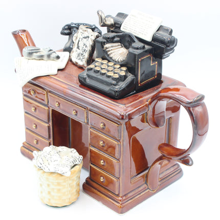A ceramic novelty teapot shaped like a brown wooden desk with drawers, topped with a black typewriter, telephone, and papers. A small waste paper basket sits beside it on a white background.