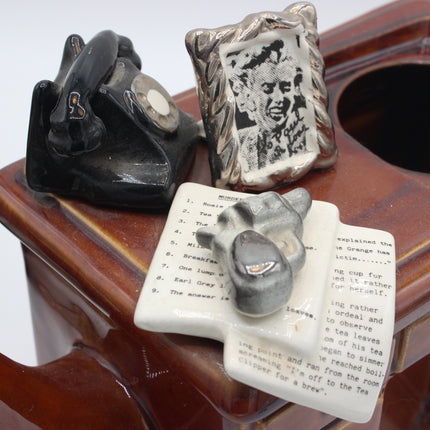A close-up of a brown ceramic teapot with three small hand-painted ceramic figurines on its lid: a black telephone, a silver-and-black portrait print, and a white book with a gray teapot resting on it.