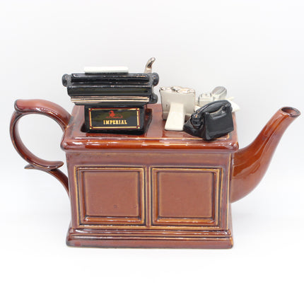 A brown ceramic teapot shaped like a cabinet, decorated with miniature toy office items including a black typewriter, a black rotary telephone, and a small silver bell, set against a plain white background.
