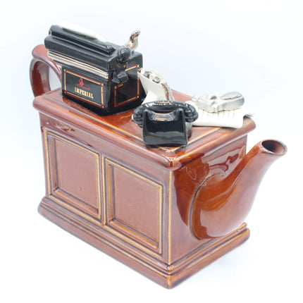A novelty ceramic teapot shaped like a brown wooden desk, featuring a black typewriter, a black rotary telephone, and a small silver skull decoration on top, set against a plain white background.