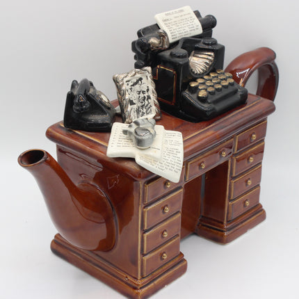 A brown ceramic teapot shaped like a vintage desk with drawers, topped with miniature black typewriter, telephone, and open letter props, photographed on a light gray studio background.