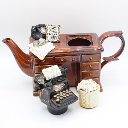A ceramic figurine depicting a brown teapot-shaped desk with drawers, topped with a black rotary telephone and papers, with a black typewriter in front and a small cream-colored basket on the right, all on a white background.