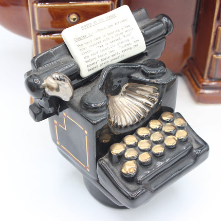 A decorative ceramic typewriter figurine in glossy black with gold-toned keys and a small white paper sheet on the platen. The figurine is photographed close-up on a white surface with a blurred brown ceramic background.