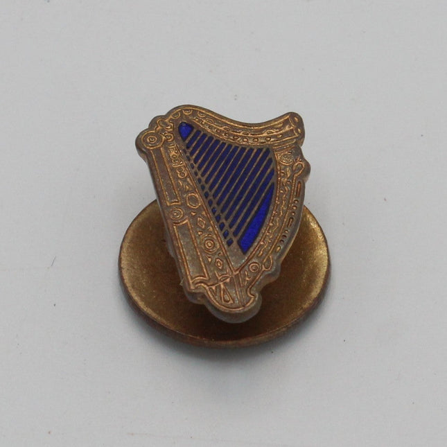 A small decorative guinness pin shaped like a harp, featuring a gold-toned metal body with a blue striped center, mounted on a round gold-toned base against a plain light background.