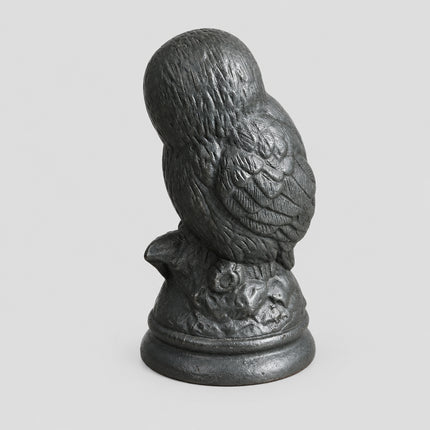 A dark slate owl figurine standing on a round base, photographed on a brown cardboard surface with a dark gray fabric background.