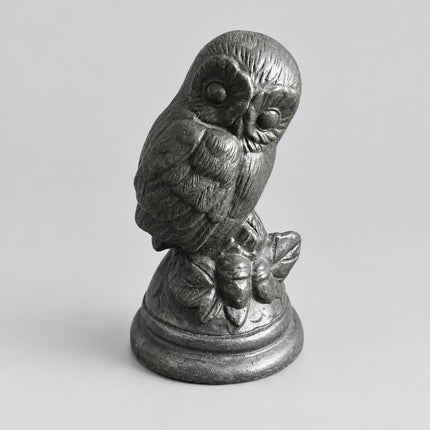 A small carved owl figurine in a dark gray stone or resin finish, perched on a leafy branch atop a round base, set on a brown surface with a gray upholstered chair in the background.