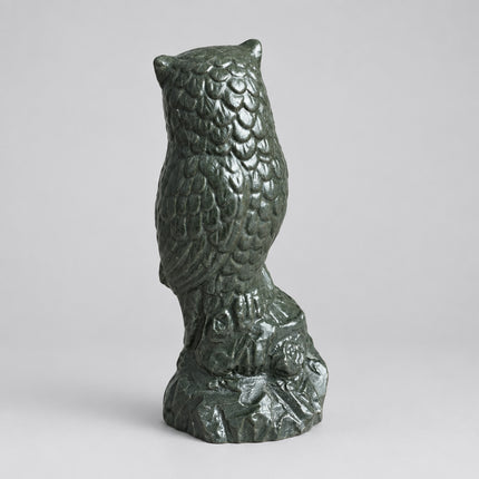 A small owl figurine or candle holder with textured feather details, standing on a tree-branch-like base, photographed on a brown surface with a gray upholstered chair in the background.