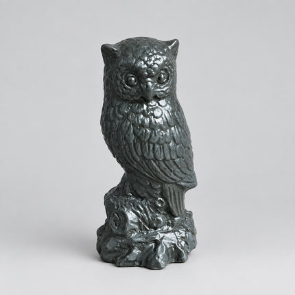 A small owl figurine in a dark metallic gray finish, perched on a textured base, set on a brown cardboard surface with a gray fabric background.