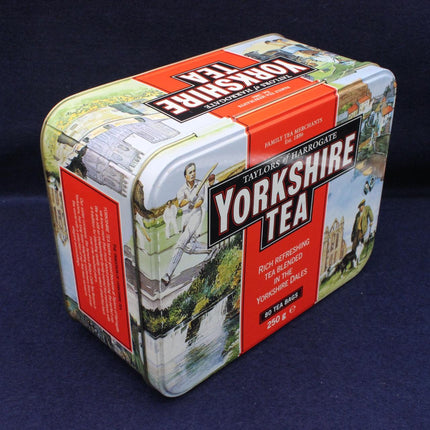 A vintage-style metal tin of Taylor's of Harrogate Yorkshire Tea, featuring illustrated scenes of Yorkshire Dales and a red label with white and black text, photographed on a dark blue background.