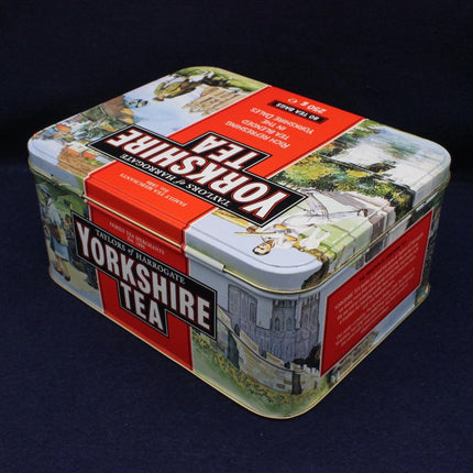 A square metal tin of Yorkshire Tea on a dark navy background. The tin features vintage-style illustrations of Yorkshire landscapes and buildings, with prominent red and white labels and the brand name 'Yorkshire Tea' in bold white text.