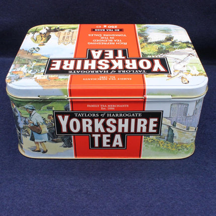 A vintage-style rectangular metal tea tin for Taylor's of Harrogate Yorkshire Tea, featuring a red central band with white lettering and illustrated scenes on the sides, photographed on a dark blue background.