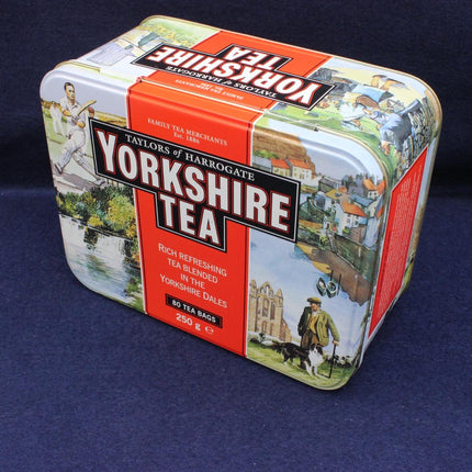 A vintage metal tin of Yorkshire Tea, featuring the brand name 'Taylor's of Harrogate' and 'Yorkshire Tea' in bold white text on a red background, surrounded by illustrations of the Yorkshire Dales landscape. The tin is photographed on a dark blue textured fabric background.