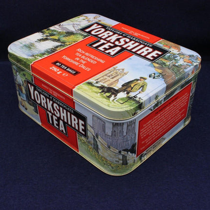 A square metal tea tin with vintage-style illustrated panels and large 'Yorkshire Tea' branding on the lid and side, photographed on a dark navy background.
