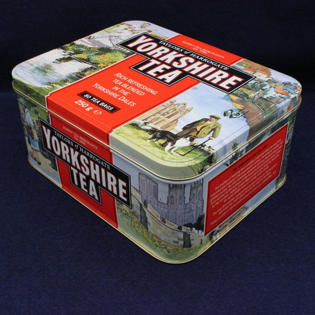 A square metal tea tin with vintage-style illustrated panels and large 'Yorkshire Tea' branding on the lid and side, photographed on a dark navy background.